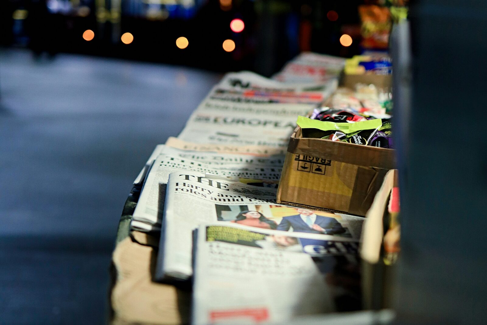 Ein Zeitungsstand bei Nacht, viele Titel nebeneinander, dahinter unscharfe Straßenlichter.
